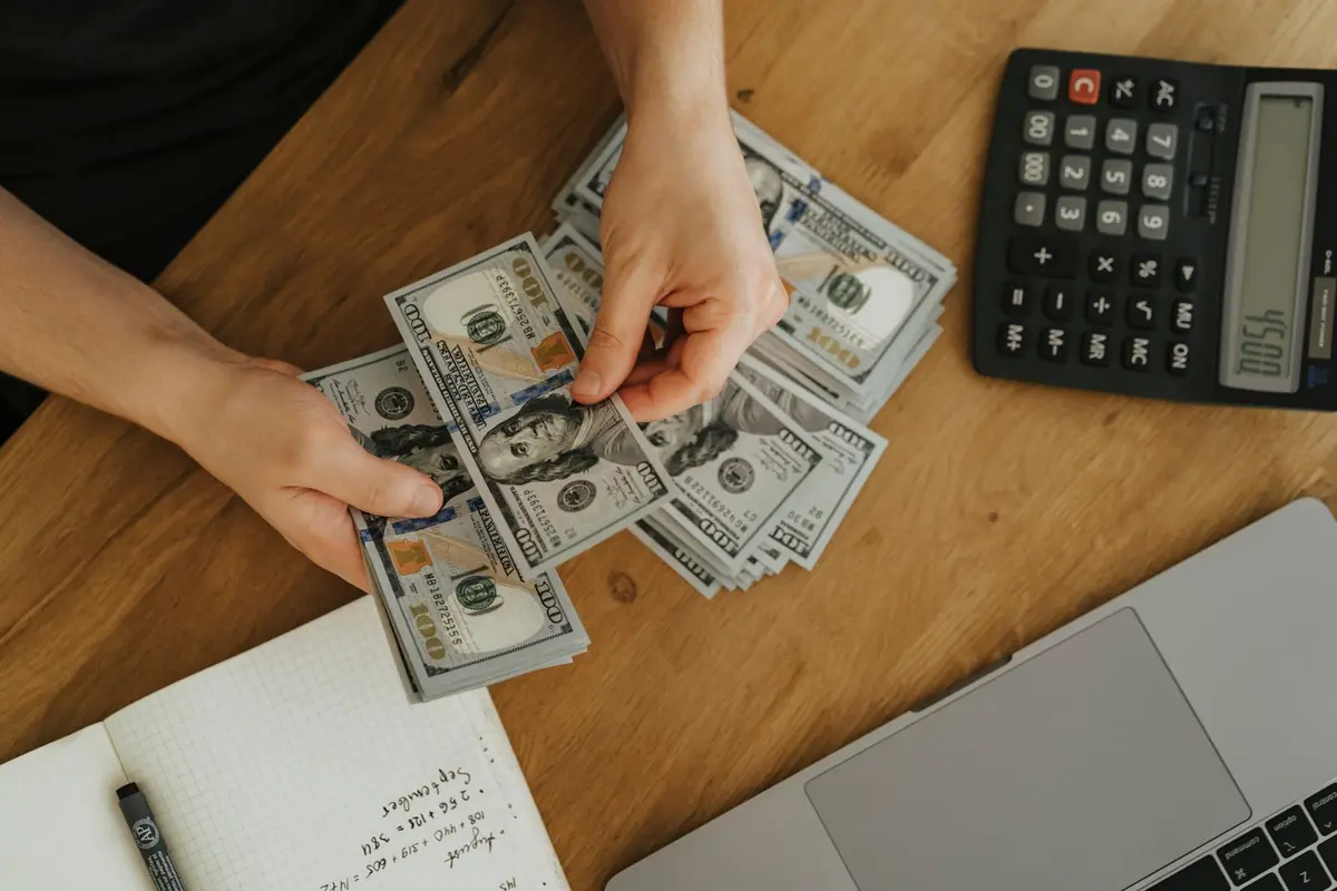 Hands counting hundred dollar bills on a wooden table beside a calculator and a handwritten budget notebook