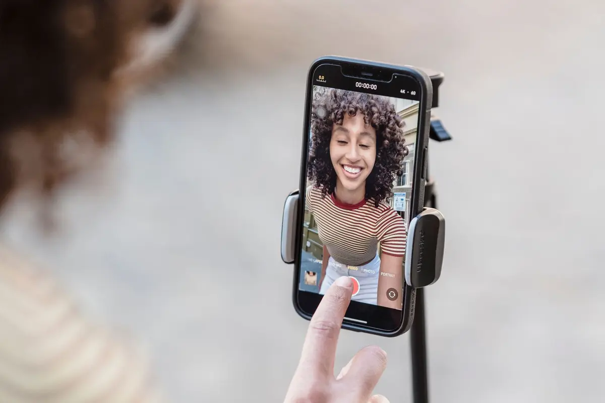 Over the shoulder view of a young woman tapping the record button on her phone, mounted on a tripod outdoors