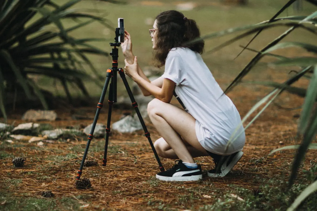 A young content creator crouches on a rural path, setting up her phone on a tripod to film a video outdoors