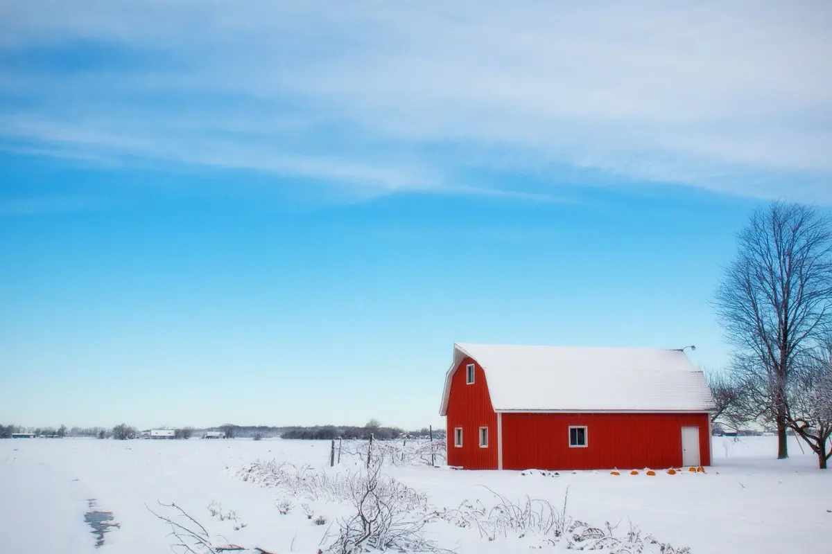 Red barn with a snow covered roof in a quiet snowy farm field under a blue winter sky