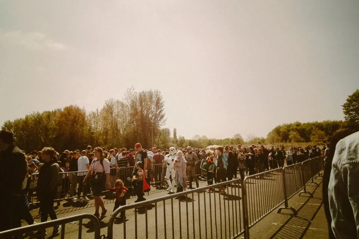 A long line of visitors waiting between crowd barriers at an outdoor event entrance on a sunny day