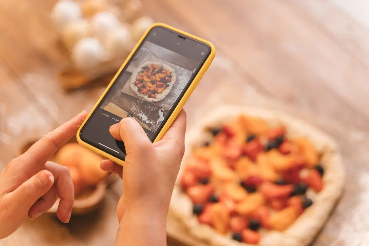Hands photographing a fresh fruit galette with a smartphone on a warm wooden farm table