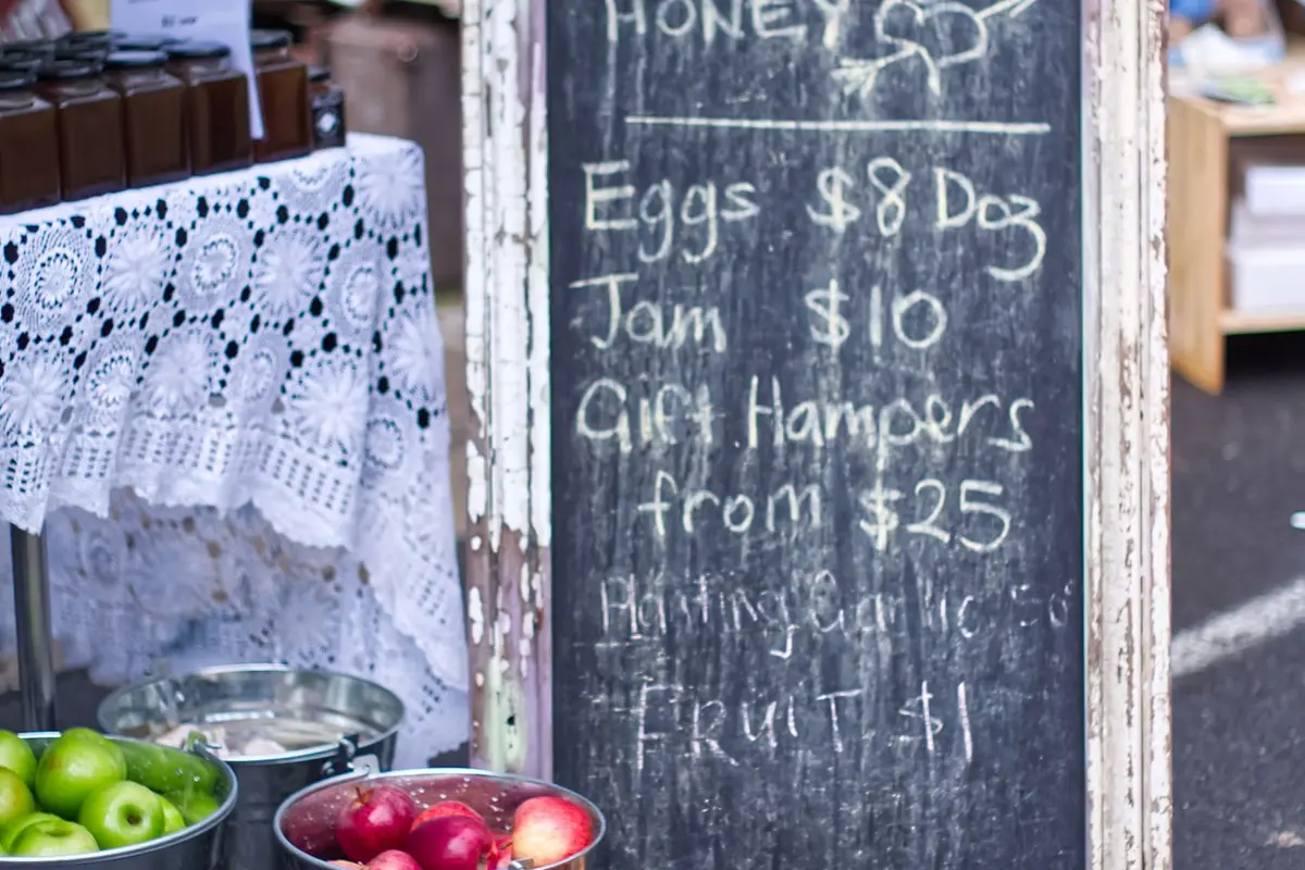 A weathered chalkboard sign at a farm stand listing prices for eggs, jam, and gift hampers beside buckets of apples