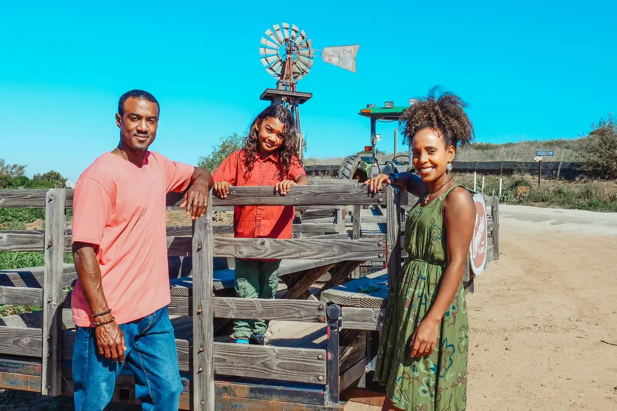 A smiling family of three gathered around a wooden hayride wagon at a sunny farm