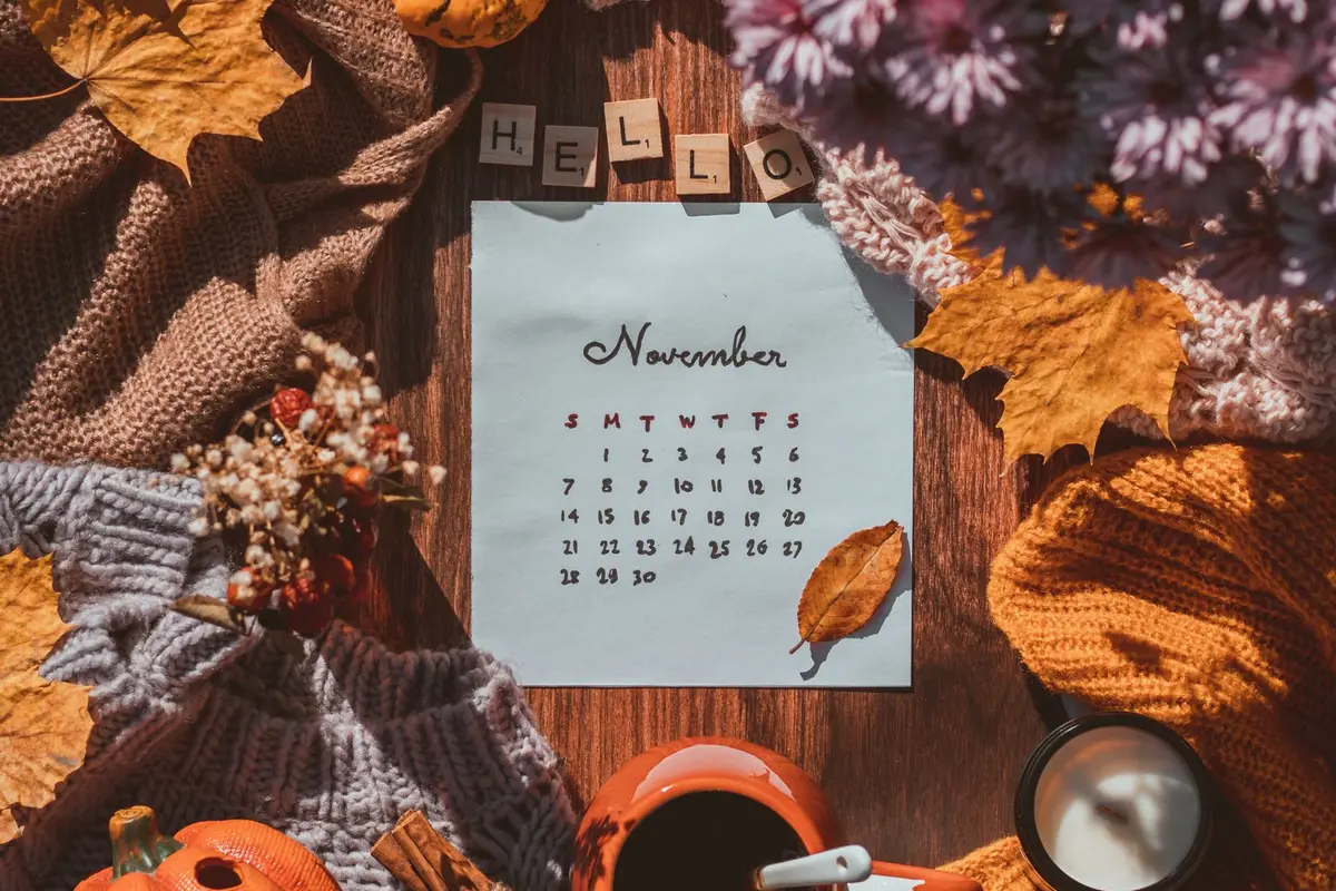 A hand drawn calendar page on a wooden table surrounded by autumn leaves, knit blankets, and a cup of coffee