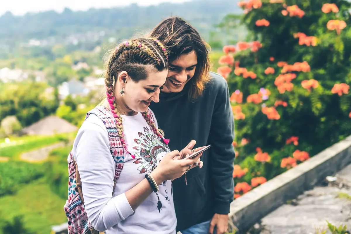 A smiling couple outdoors reading a message together on a phone, with green hills and flowers behind them