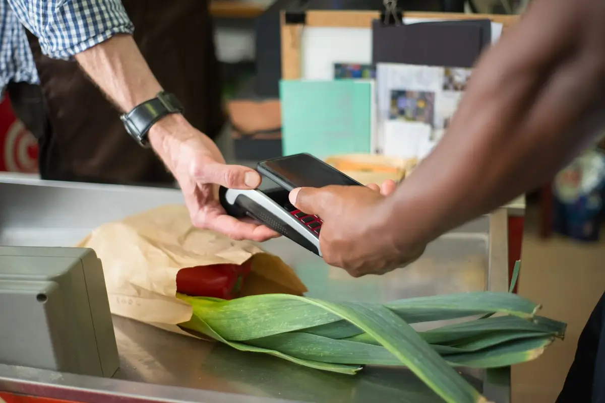A customer tapping their phone to pay at a market checkout with fresh produce on the counter