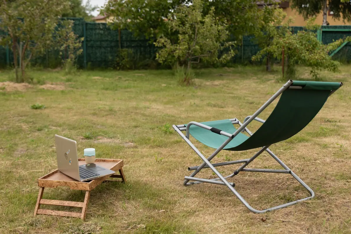 Laptop open on a small wooden table in a grassy backyard, with a folding chair and fruit trees behind it